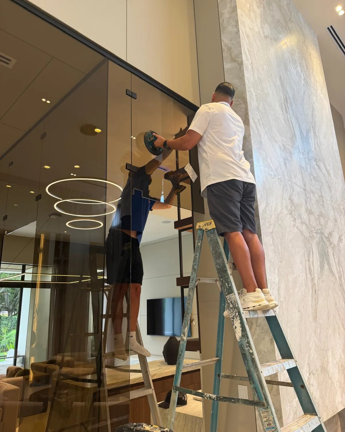 Two Galaxy Glass installers on ladders fitting a tall glass panel beside a marble wall
